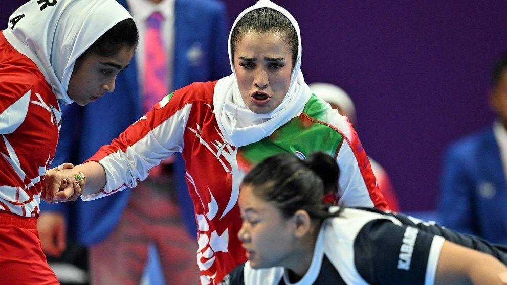 Two women, wearing white head scarfs and red and white kits, are focused on another woman, who is wearing a black and white kit. 