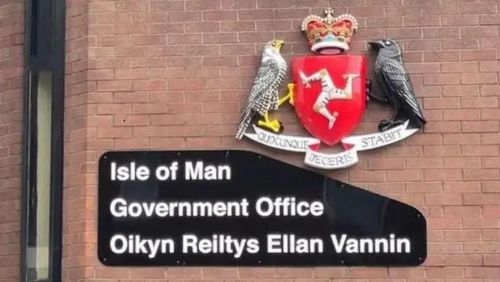 A sign on a red brick wall reads 'Isle of Man Government Office' in both English and Manx. The sign features white lettering on a black background, and sit next to the government crest.