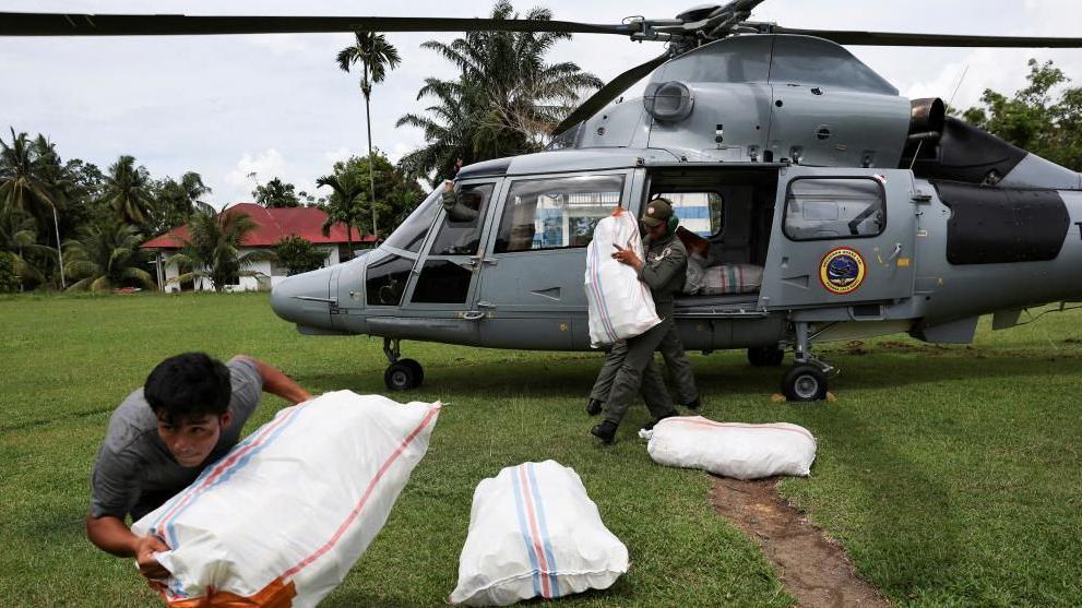 A young man carries a sack of aid from a helicopter across a green field.