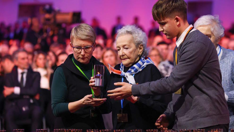 An elderly woman who survived the Birkenau death camp is helped by a younger woman and a young man to place a candle