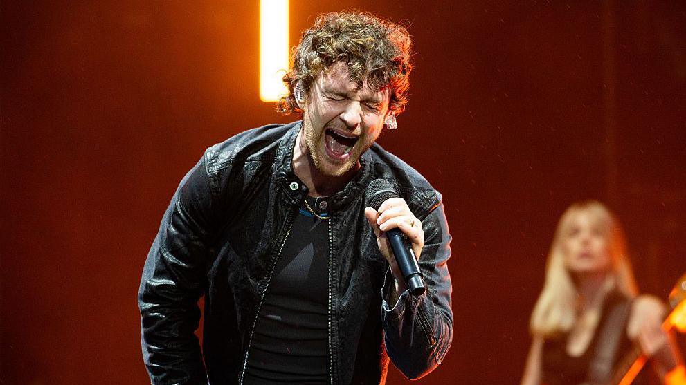 Tom Grennan is on stage singing into a microphone wearing a leather jacket