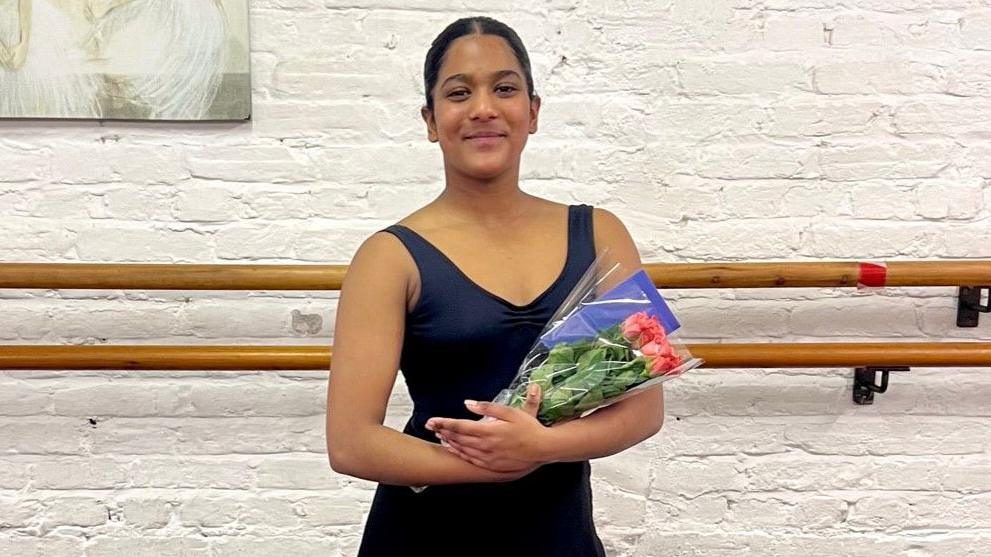 Jemima wearing a black leotard and holding a bunch of flowers. She is smiling at the camera. She is in a ballet studio with a bar behind her and a white brick wall. 