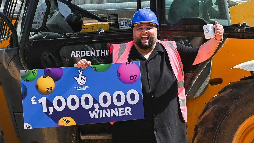 Adam Lopez, a man who is standing outside in front of a forklift. He is wearing a black button up shirt, pink hi-vis jacket and blue hard hat. He is looking directly at the camera and is smiling. He is also holding a blue National Lottery sign which says £1,000,000 winner in white writing.