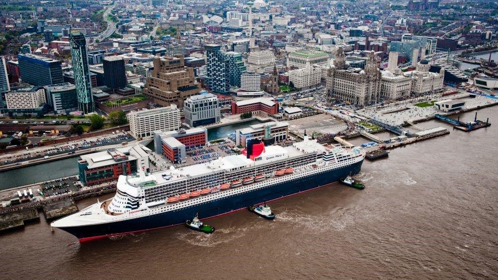 An aerial view of the Queen Mary 2 ocean liner, with white decks and a blue and red hull, berthed on the River Mersey with three small green barges pushing against its port side.
