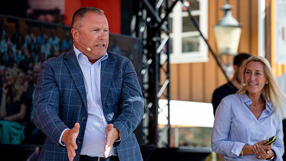 A man in a checked-blue jacket and light blue shirt stretches his hands out as he talks, while a woman looks on