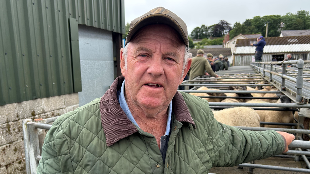 Llun o ffermwr yn ei 70au mewn mart amaethyddol gyda chorlannau defaid tu ôl iddo. Mae e'n gwisgo cap ar ei ben a chot werdd