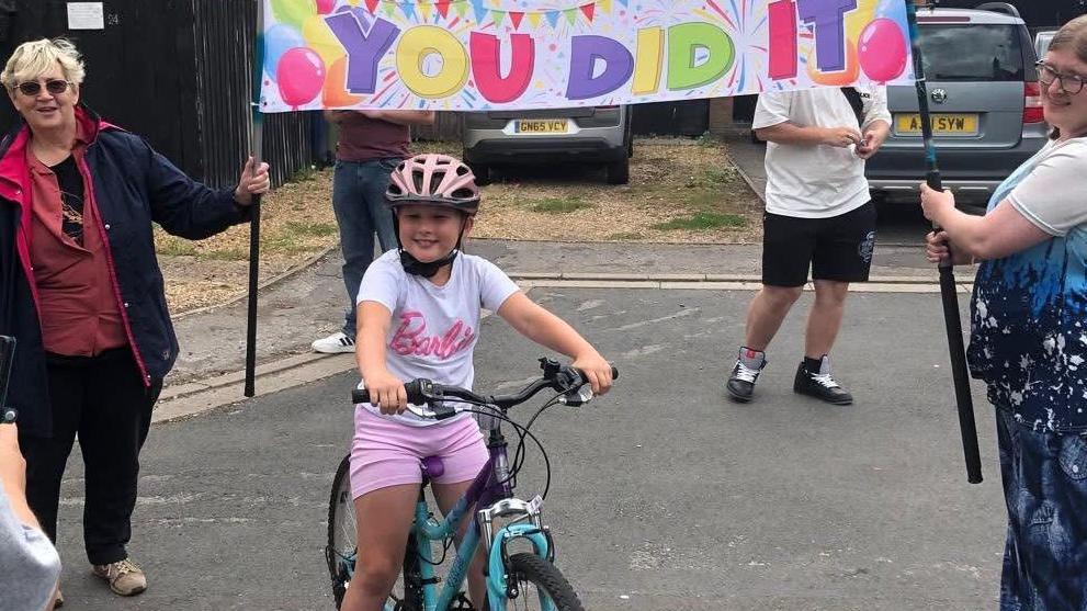 Evie is sitting on a blue bike while wearing a pink helmet, pink shorts and white top with 'Barbie' written in pink in the centre. She is smiling as people stand around her on a street. Behind her two women are holding a white sign that reads 'You did it' with balloons and confetti. 