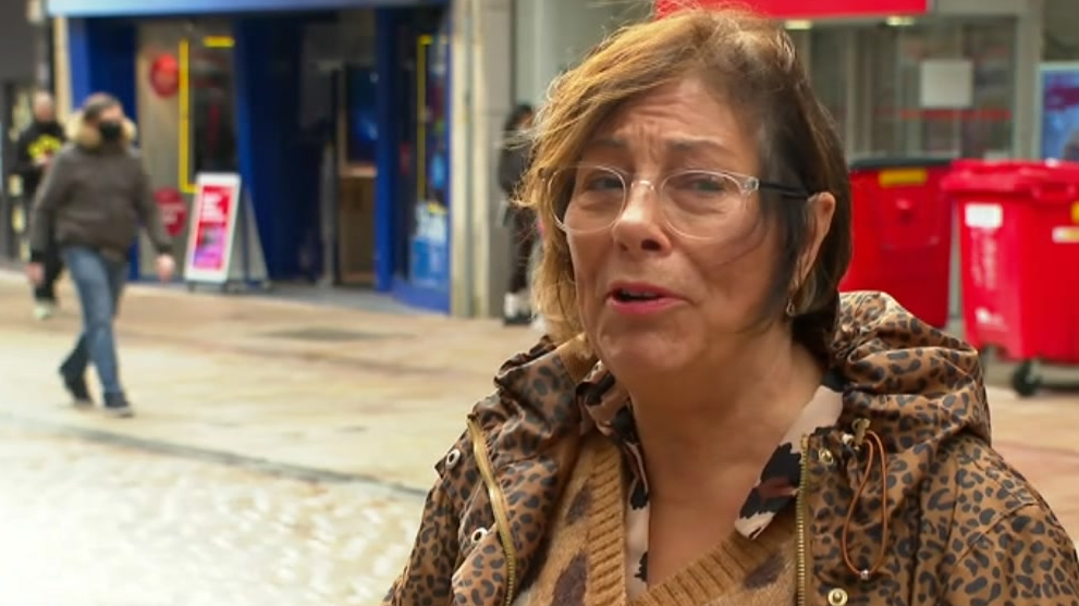 Julie is stood in a street in the city wearing a patterned coat and brown jumper and glasses. She has short brown hair with pink lipstick on.
