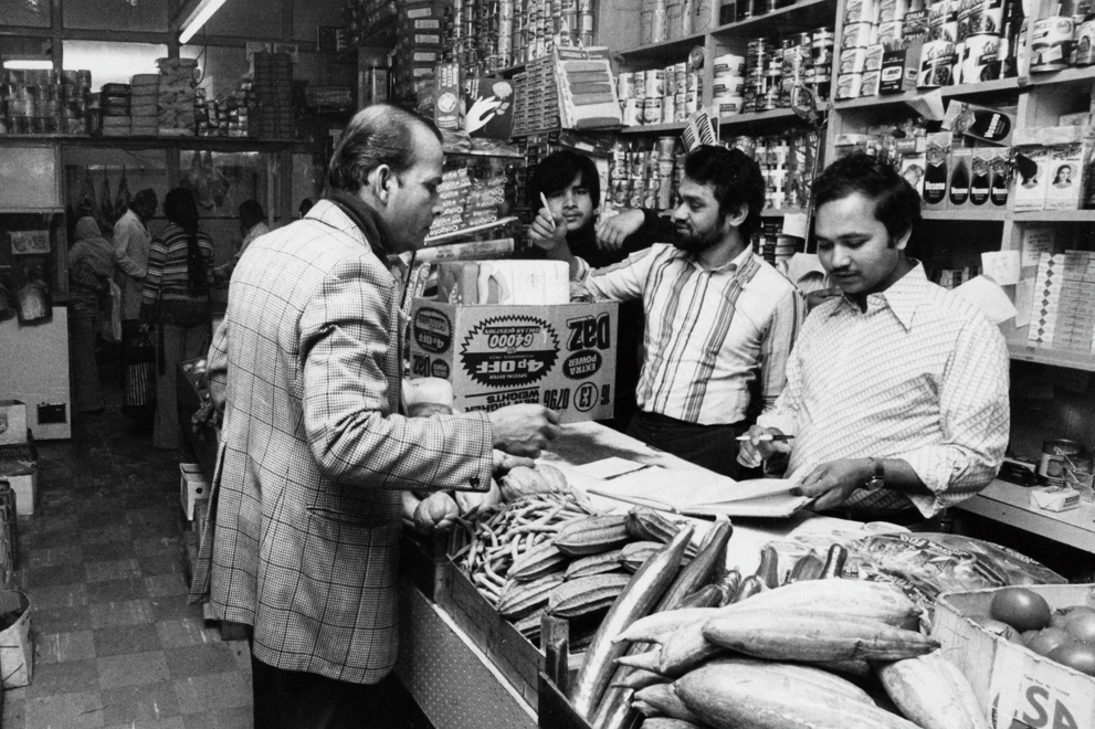 Black and white photo of shopkeeper Komor Uddin serving a customer at his grocer's shop, the Taj Stores in Brick Lane