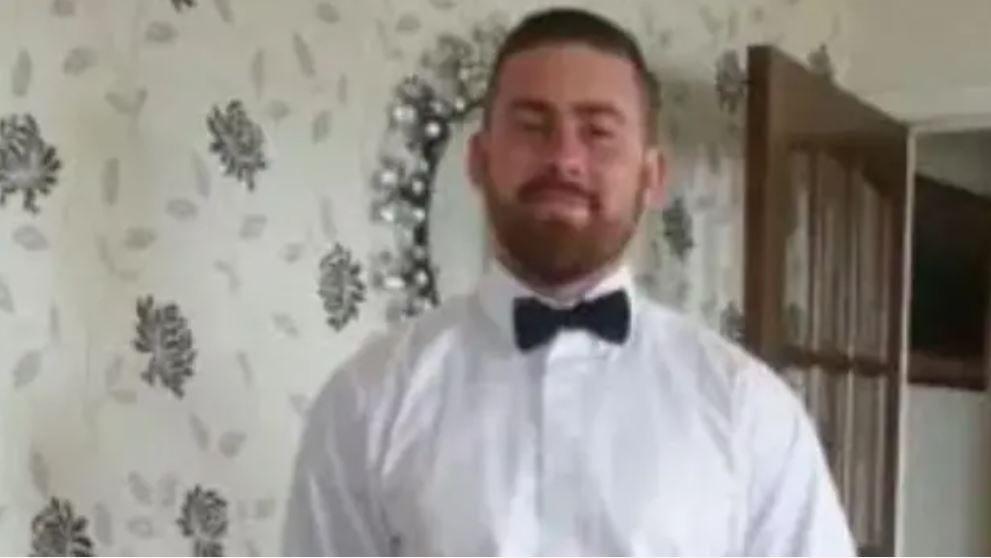 A bearded man with short brown hair, a white shirt and a black bowtie smiling at the camera in front of a wall.