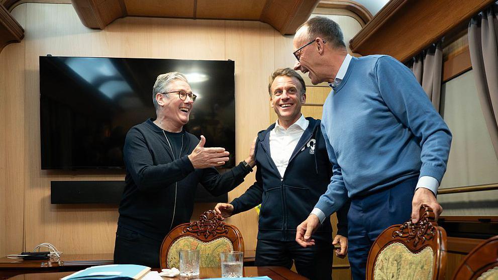 Keir Starmer smiles and gestures with his hands while in conversation with Emmanuel Macron and Friederich Merz, both of whom are laughing. Starmer and Macron are wearing dark zip up hoodies while Merz is wearing a light blue sweater.