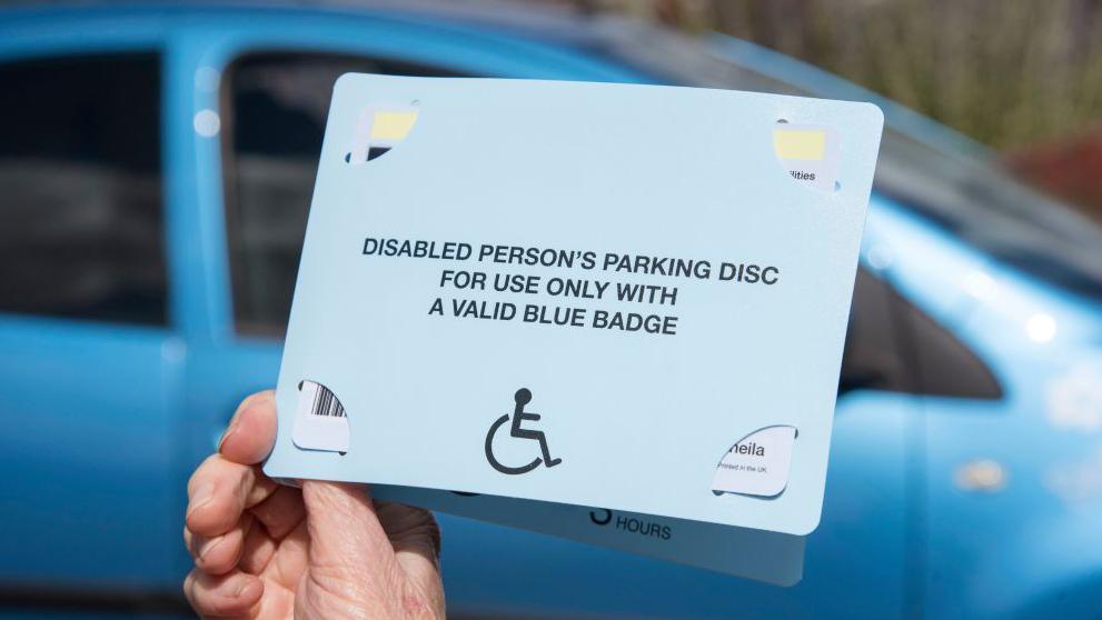 A woman is holding a blue badge in front of a blue car. The badge has writing on the front which reads, DISABLED PERSON'S PARKING DISC FOR USE ONLY WITH VALID BLUE BADGE.