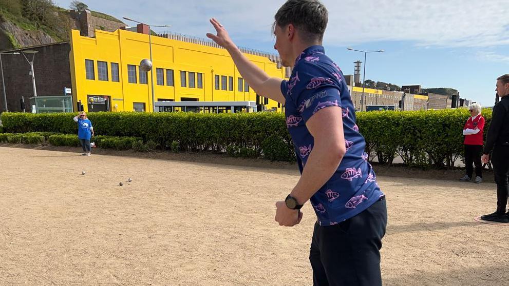 A man wearing a navy blue shirt with pink fish on it throws a petanque ball during a game. A building with bright yellow walls is in the background.