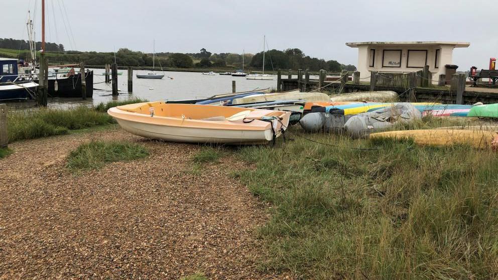 The river Deben from the quayside. It's a slightly grey day and the picture is looking out onto the river. There is a small shingle river bank with boats in the foreground and then a shelter. There are some boats on the river too. 