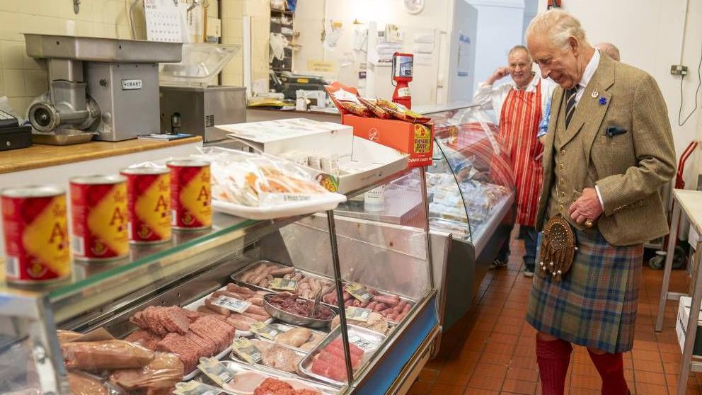 King Charles wearing a kilt standing in front of a glass counter with raw meat enclosed.