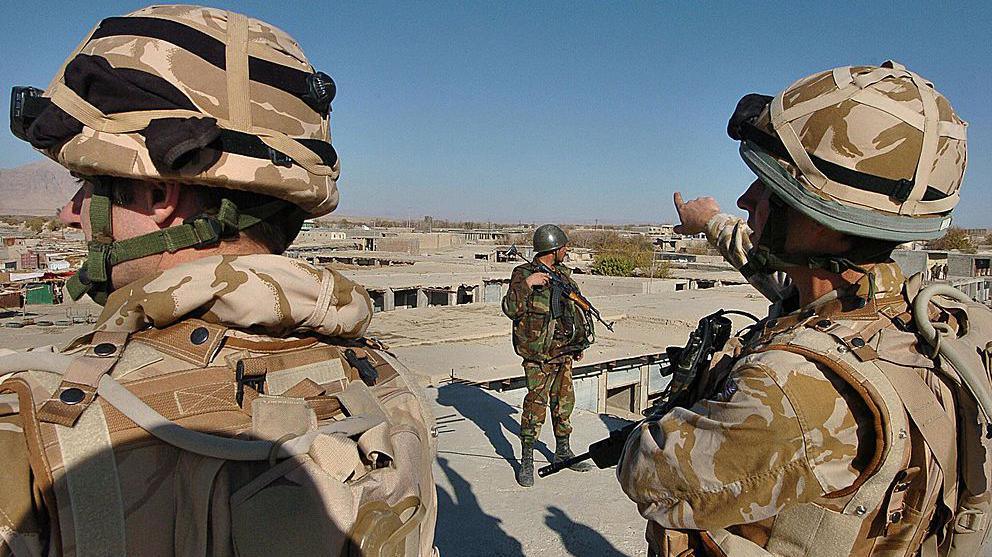 Two British Army soldiers wearing desert fatigues stand close to the camera in an urban landscape in Afghanistan. An Afghan National Army soldier stands, framed by the British troops.