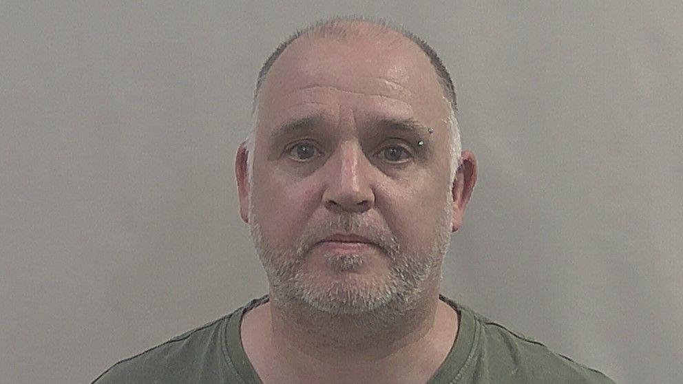 A police headshot of Stacie John Farrar. He has short/ balding dark-coloured hair on his head and short grey facial hair. He is wearing a khaki green t-shirt. 