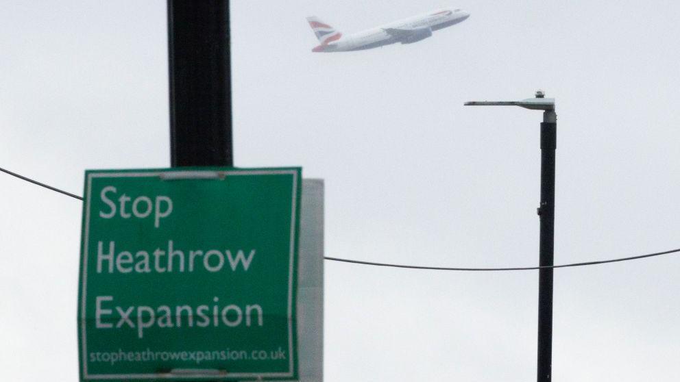 A green sign on a lamppost that reads, in white lettering: "Stop Heathrow Expansion". The sky in the background is white as though its winter and there is a plane in it.