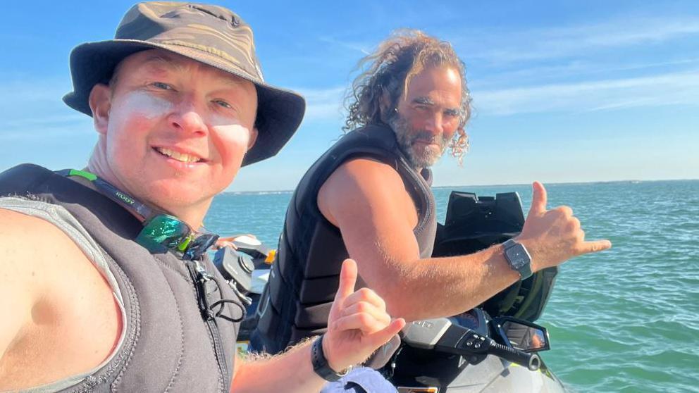 Ben Felton (left) and Nick Woods sitting on a silver water scooter on the sea - which is a greenish, blue colour. The sky is blue with a couple of lines of very thin, wispy clouds. Mr Felton is taking the selfie image. He is wearing a brown bucket hat and a grey lifejacket. He also has a pair of sunglasses resting under his chin and he has a line of white sunblock going horizontally across each cheek. Mr Woods is also wearing a grey lifejacket. He has shoulder-length dark, curly hair and a greying beard