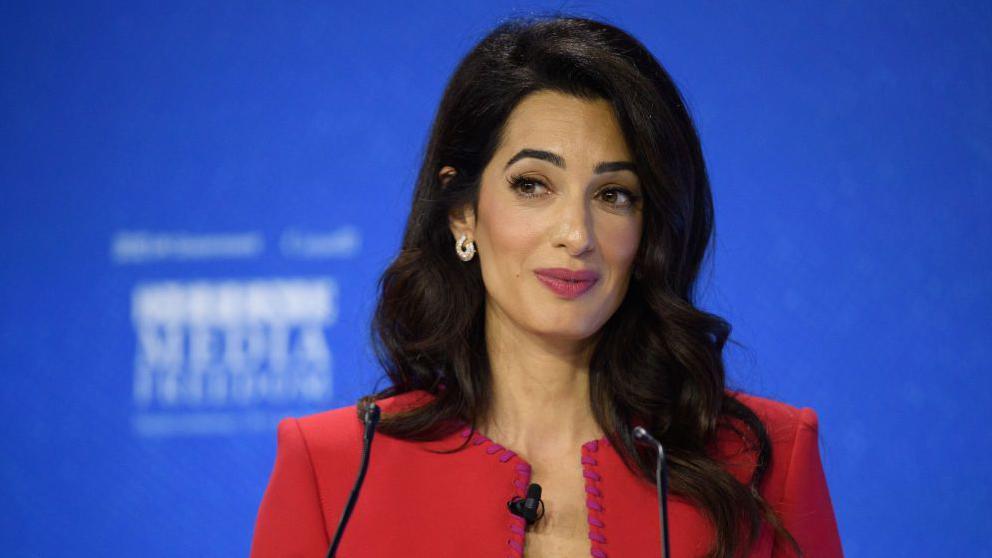 Amal Clooney wears a smart red jacket, and stands at a podium, possibly giving a speech. She has long dark hair which comes past her shoulders.