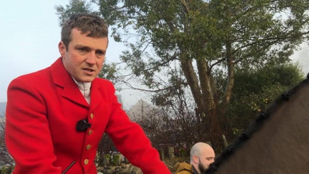 A man sat atop a horse is on the left of the shot looking just off camera. He is wearing traditional red hunting outfit. Behind him is a large tree.