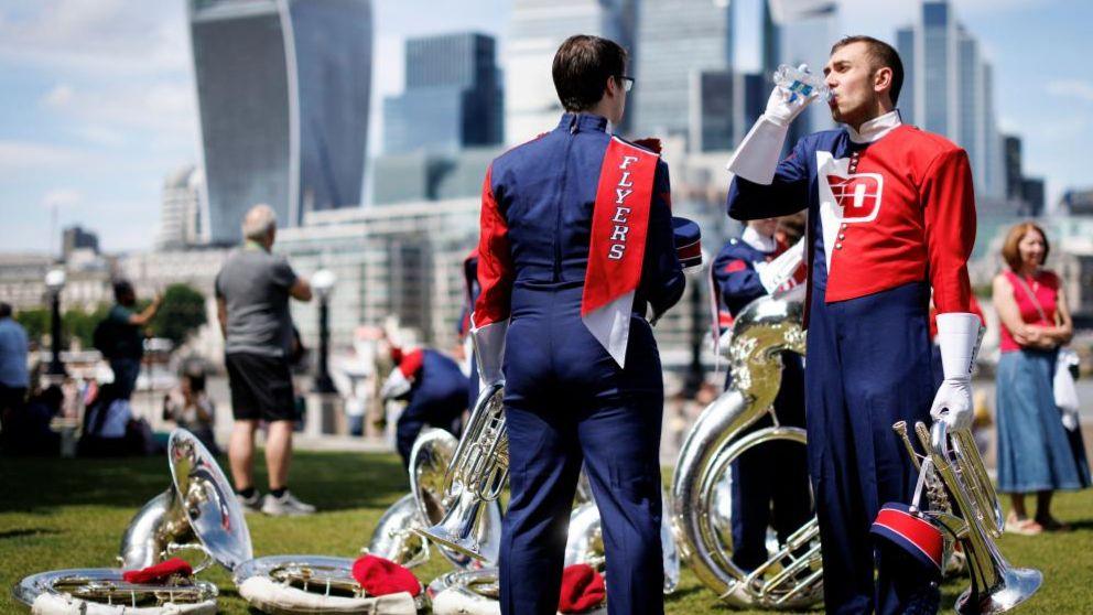 Two members of the Pride of Dayton Marching Band drink water as they take a break from performing at Potters Field Park during a hot day in London. They are dressed in band uniforms of blue, white and red. They stand against a background of brass instruments lying on grass.