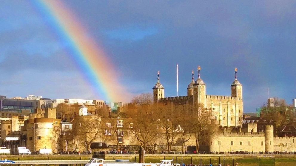 A multicoloured rainbow over the Tower of London, a castle-like structure. The sky is still grey from the rain.