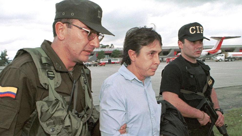 Two officers in khaki uniforms escort Ochoa, wearing a light blue buttoned shirt, across an airport tarmac