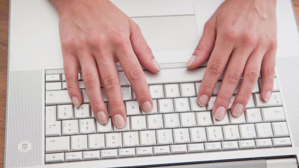 Stock image showing a grey laptop keyboard. A woman's hands are resting on the keys as if about to type.