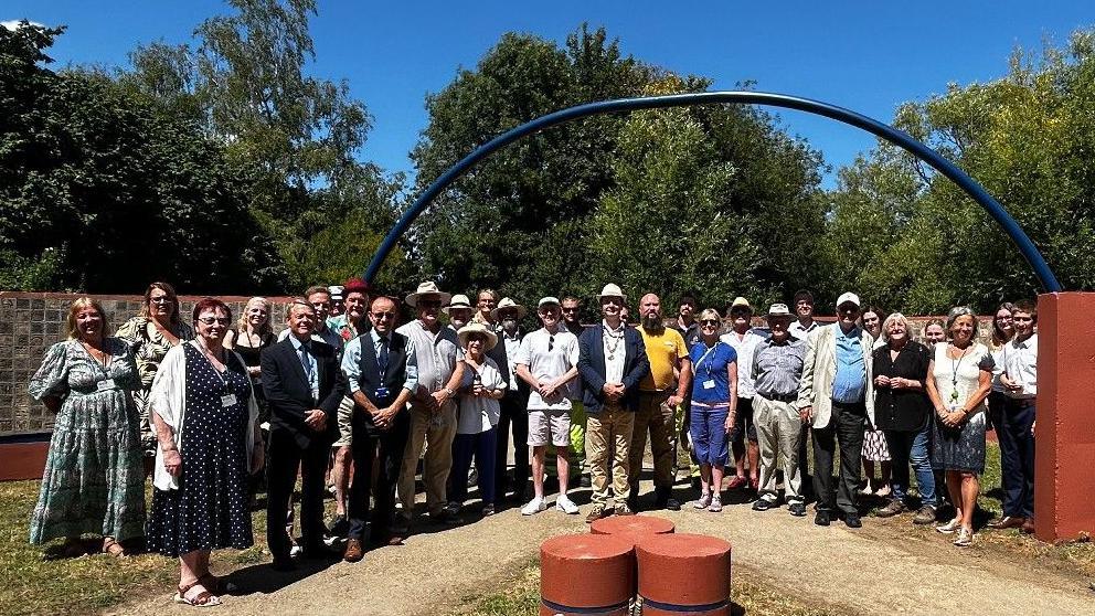 A large group of people on a sunny day, standing in front of the wall on the footpath. All are looking at the camera, the wall and trees behind them. Connecting the two halves of the wall over the path is a long arched blue bar.