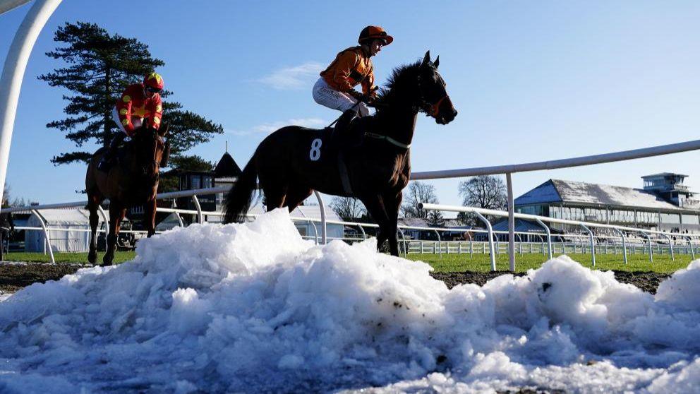 Two riders on horses at Lingfield Racecourse in Surrey, with heaps of snow in the foreground and bright sunshine elsewhere