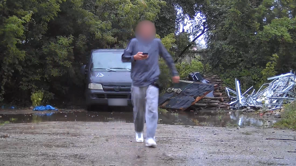 A surveillance photo of a male teenager wearing grey sweat pants and shirt and with his face blurred, carrying a mobile phone. He is walking away from a grey van parked on hard surface in a wooded area. 