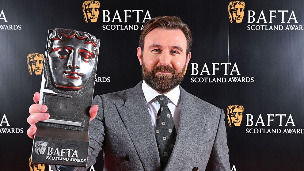 A bearded man in a grey jacket holds up a Bafta Scotland trophy