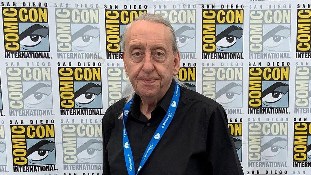 A man in a black shirt with long silver hair stands in front of a San Diego Comic Con sign