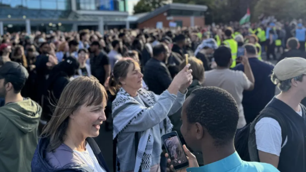 A large crowd gathered in front of a glass-fronted building which can just be seen in the background. The crowd are talking amongst themselves and holding up phones to take pictures.