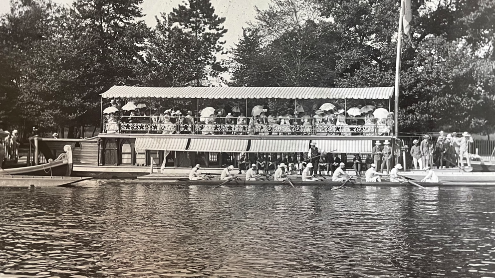 A black and white archive image of rowers passing by an Oxford college barge. A crowd of people watch them from the sides and on board the vessel. It appears to have been a sunny day.
