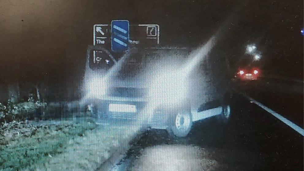 A white van sits at the side of a road, with its lights on, facing oncoming traffic.