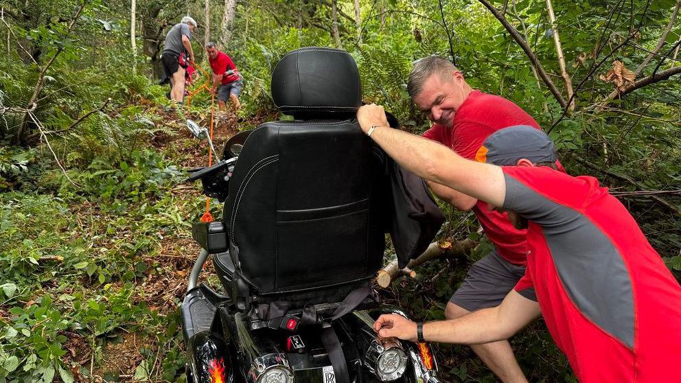The mountain rescue team trying to recover the scooter. Two men are pushing it from behind up the hill. They are hearing red and grey t-shirts. At the top of the hill two men can be seen pulling the scooter up using orange rope.