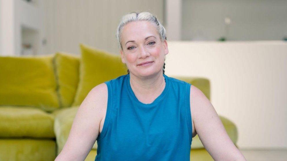 Mrs Blezard smiles at the camera as she sits on the floor in a yoga pose. Her blonde hair has been braided back behind her head. She wears a blue vest top. A green sofa can be seen behind her.