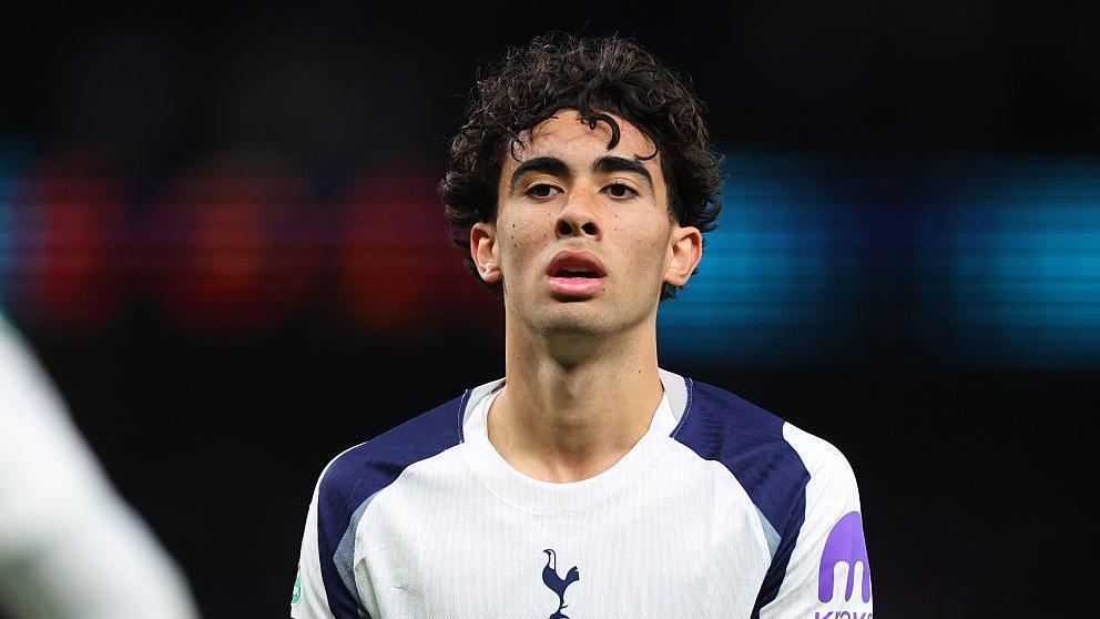 Luca Williams-Barnett has short, dark curly hair and is looking straight ahead. He is wearing a Spurs strip