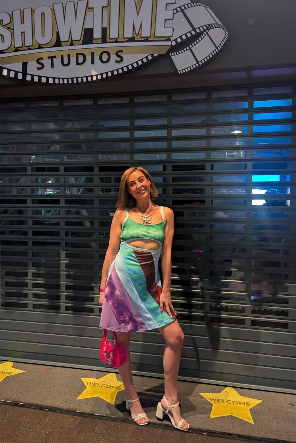 A woman wearing a multicoloured, striped skirt and green crop top, with white heels and a pink handbag. She's stood smiling, in front of a shutter, with yellow stars on the ground and a sign above her which reads: Butlin's presents showtime studios".