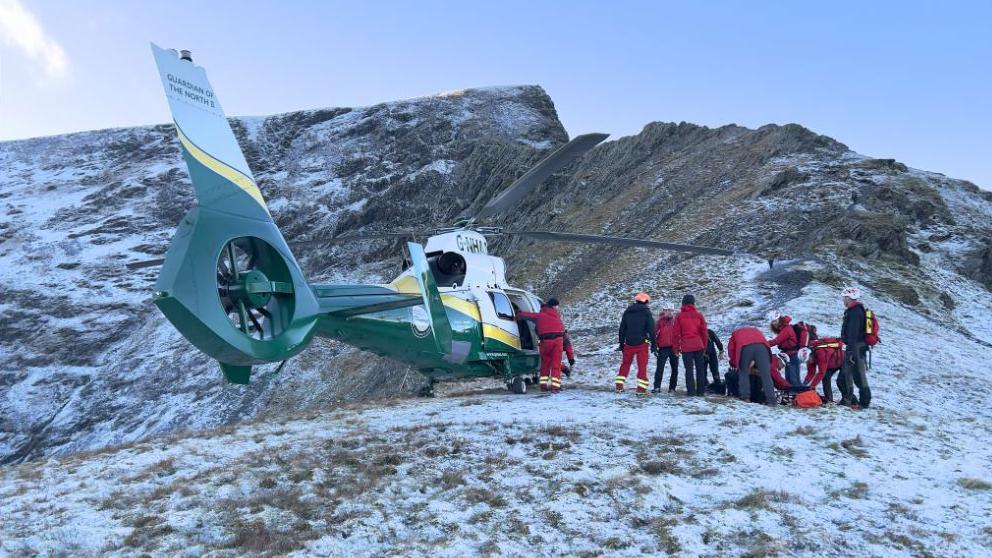 A helicopter - the air ambulance - landed on the side of a snowy mountain, with a group of mountain rescue volunteers wearing red high jackets clustered around on the right.