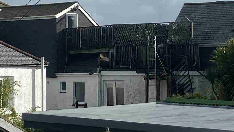 At the back of a building there is a staircase that is hanging down and a tall ladder in place where the staircase had been attached. There are palm trees nearby and the backs of other buildings.