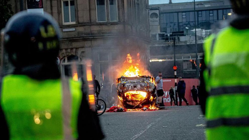 Two police officers stand at a distance from an upturned car on fire.