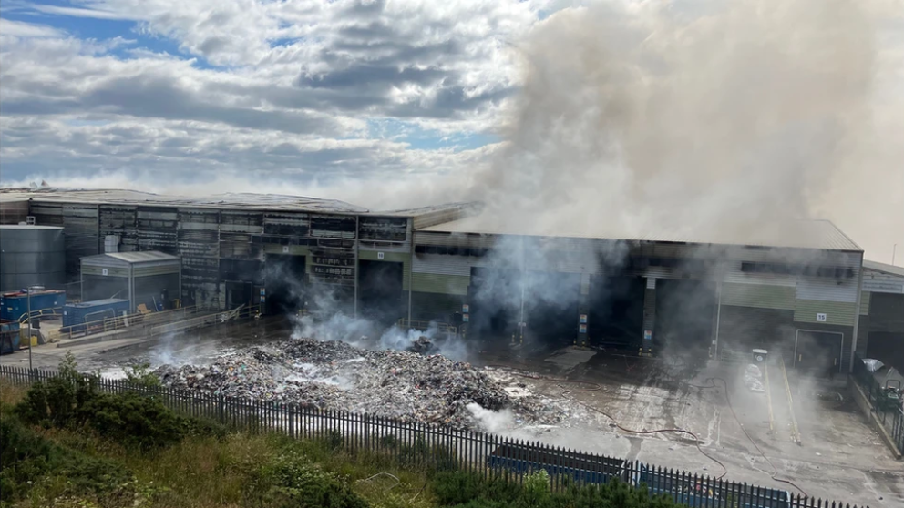 Smoke smoulders across the sites of a warehouse building housing a recycling processing facility with waste mounted up in the yard outside the building and scorch marks across the building.