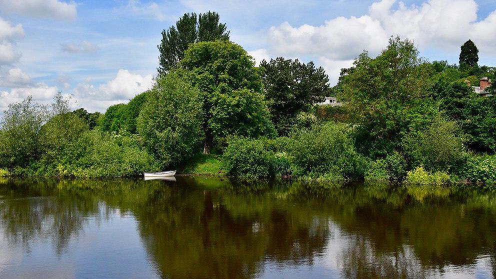 A large river in the foreground, with a small white rowing boat moored on the bank. Behind, green bushes and trees with buildings nestled in between