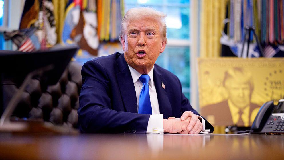 President Donald Trump speaks before introducing a $100,000 fee for H-1B visas. He is pictured in a suit and a blue tie. 