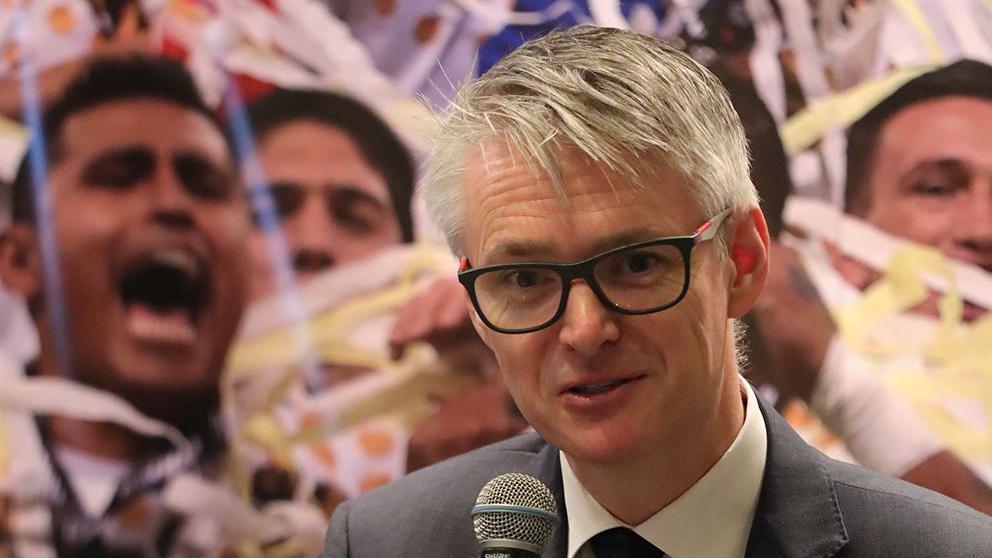 Nic Coward holding a microphone with a sports collage in the background featuring celebrating footballers and ticker-tape