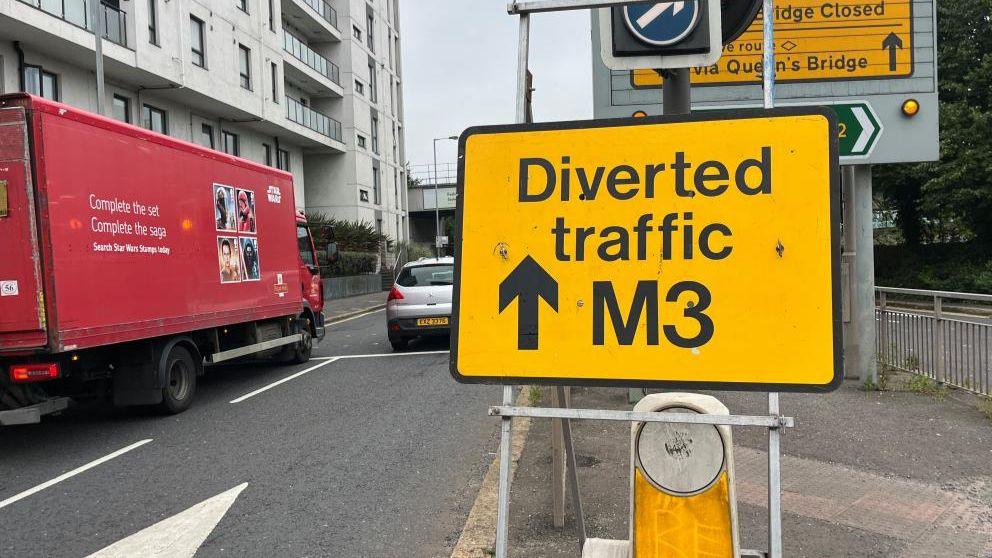 In the foreground a yellow sign with black writing says 'Diverted traffic M3'. A red lorry and a car can be seen sitting in traffic in the background.