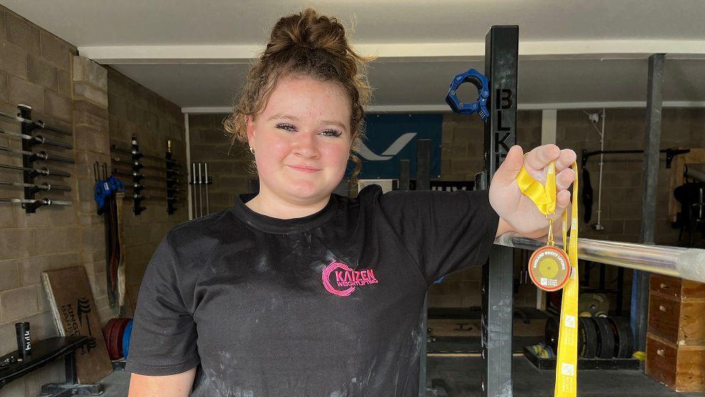 Stevie McNulty holds her medal while looking into the camera and leaning on weightlifting equipment